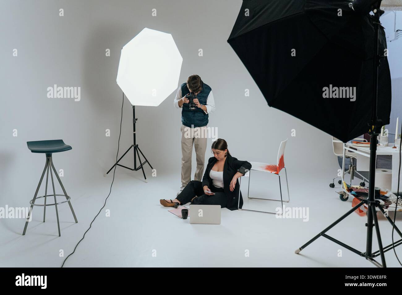 Fotografo che regola le impostazioni della fotocamera dietro le quinte in uno studio. Modello posizionato con un laptop sul pavimento, creando un bancomat dinamico per l'ambiente di lavoro Foto Stock