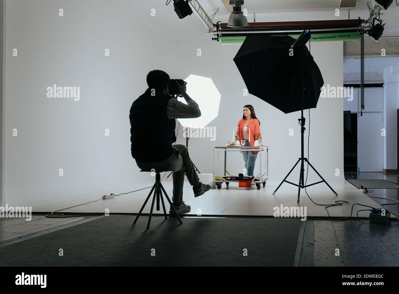 Un fotografo professionista che lavora in uno studio, cattura un modello dietro un tavolo sotto l'illuminazione dello studio, mostrando un tipico dietro lo scen Foto Stock