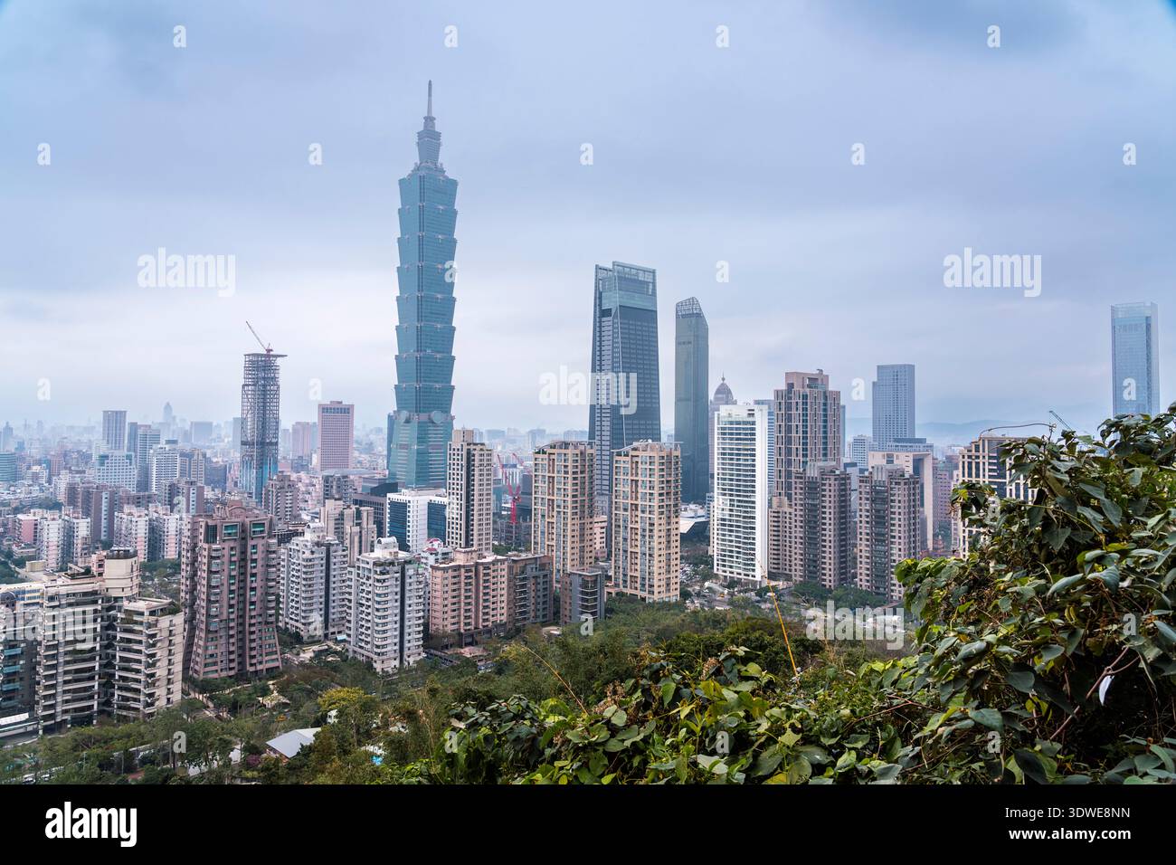 Vista sopraelevata di Taipei, Taiwan, che mostra l'iconico grattacielo Taipei 101 in mezzo al vivace paesaggio cittadino. La vegetazione lussureggiante in primo piano aggiunge un tocco naturale Foto Stock