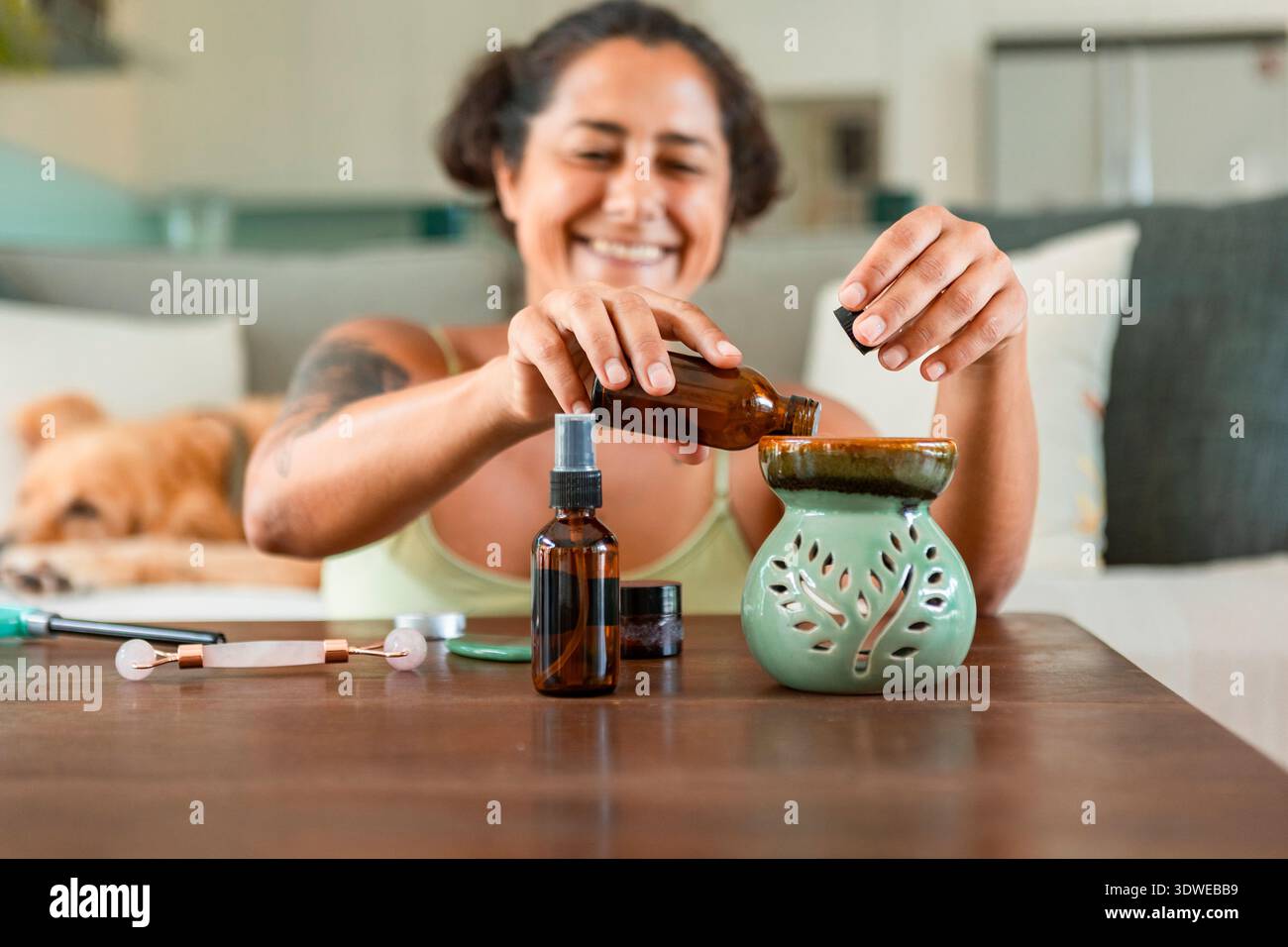 Una donna sorridente prepara una rilassante sessione di spa domestica utilizzando oli essenziali. Circondata da prodotti per la cura della pelle, crea un ambiente rilassante per se stessa. Foto Stock