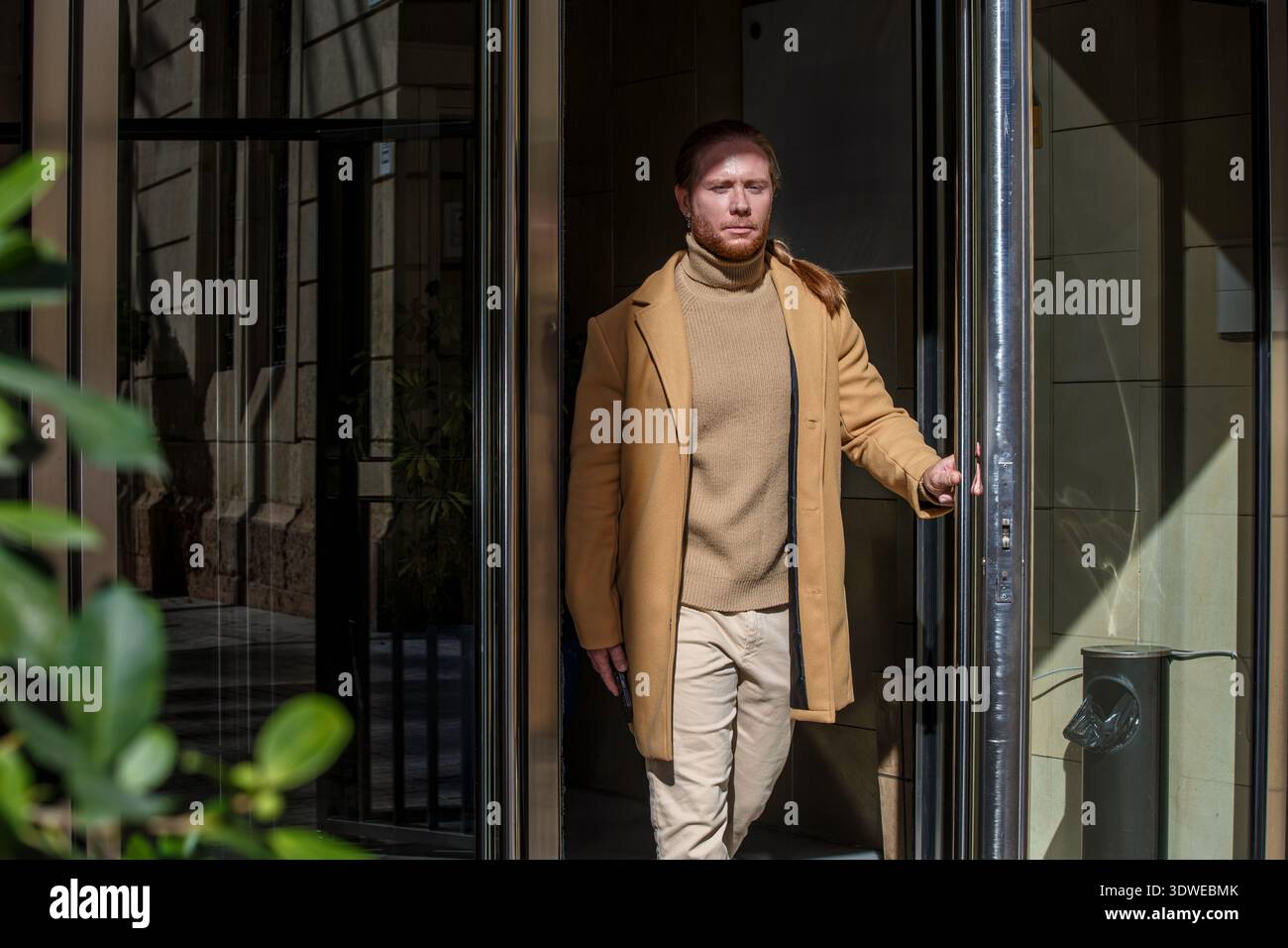 Un uomo in un informale abbigliamento da lavoro esce da un edificio, trasudando fiducia e stile. Indossa un cappotto marrone e pantaloni beige, che incarnano un mix di professionisti Foto Stock