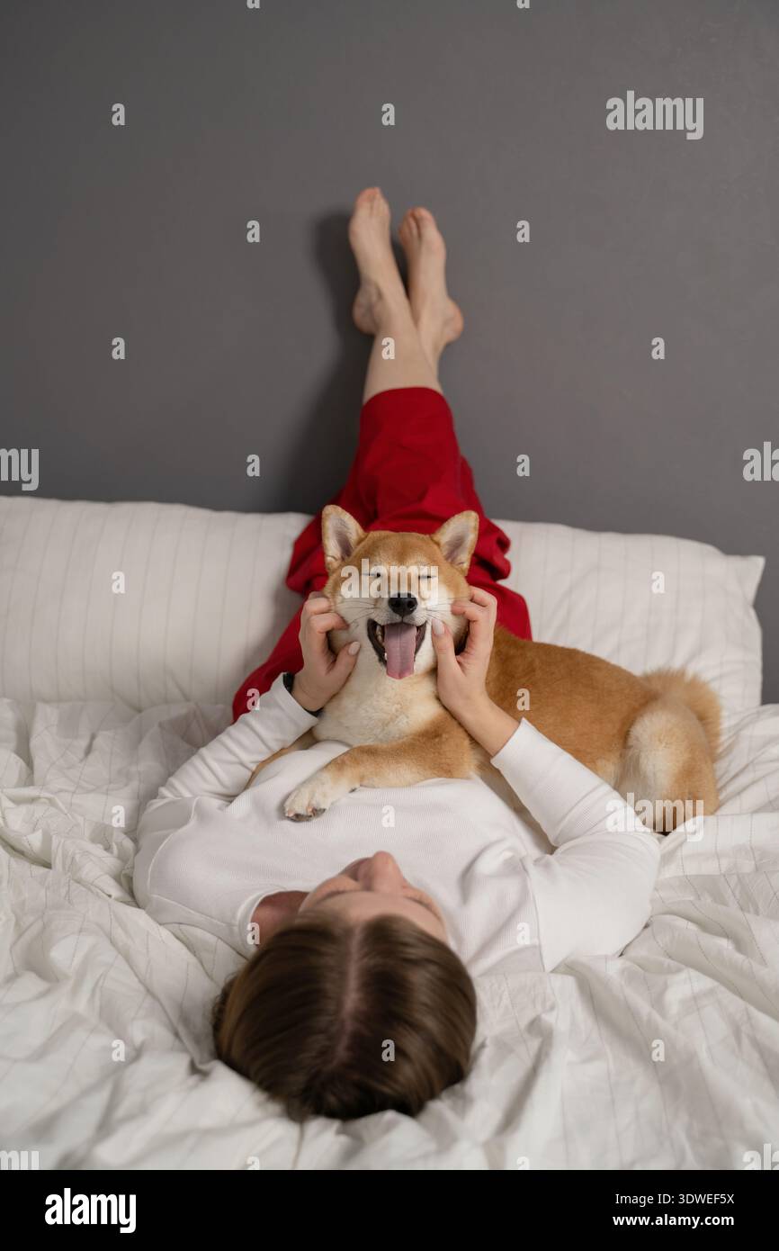 Una donna giace comodamente su un letto, tenendo giocosamente il viso del suo cane Shiba Inu. L'atmosfera rilassata e la compagnia mettono in risalto un gioioso e sereno Foto Stock