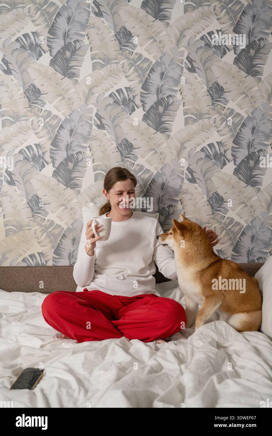 Una donna con i pantaloni rossi si siede comodamente su un letto, tenendo una tazza, mentre uno Shiba Inu si siede accanto a lei. L'ambiente e' luminoso e intimo con wallpa a motivi geometrici Foto Stock