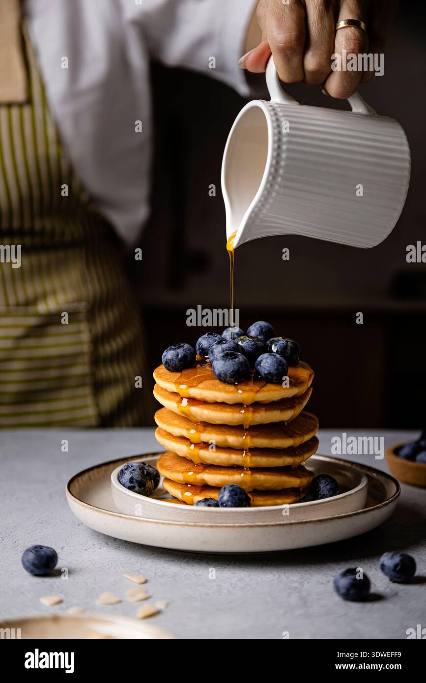 Una donna versa elegantemente lo sciroppo su una pila di soffici pancake conditi con mirtilli freschi. La deliziosa colazione è il modo migliore per iniziare la giornata Foto Stock
