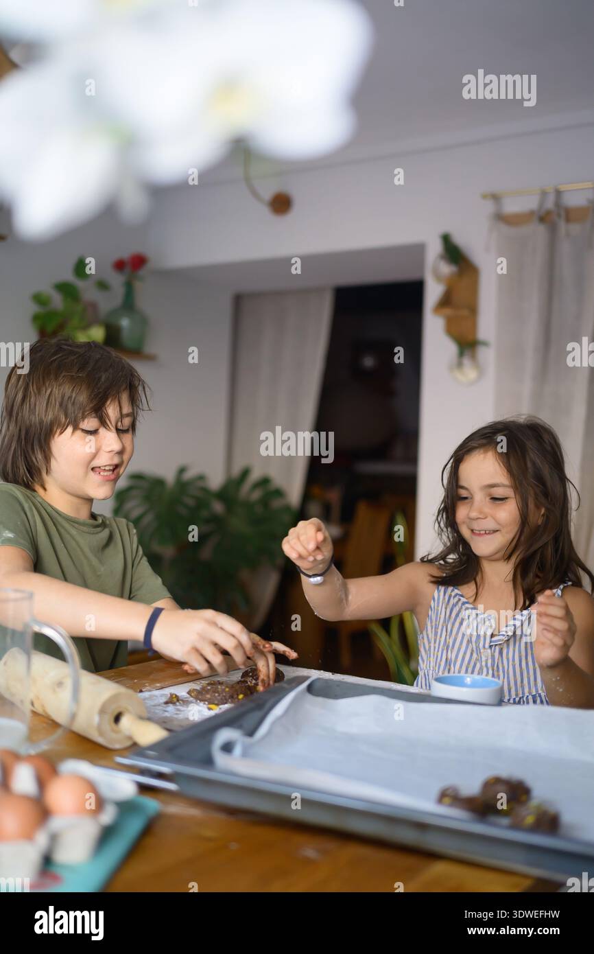 Due bambini felici si divertono con una divertente sessione di cottura in cucina, modellando e preparando l'impasto insieme ai sorrisi, circondati da ingredienti e da cuocere Foto Stock