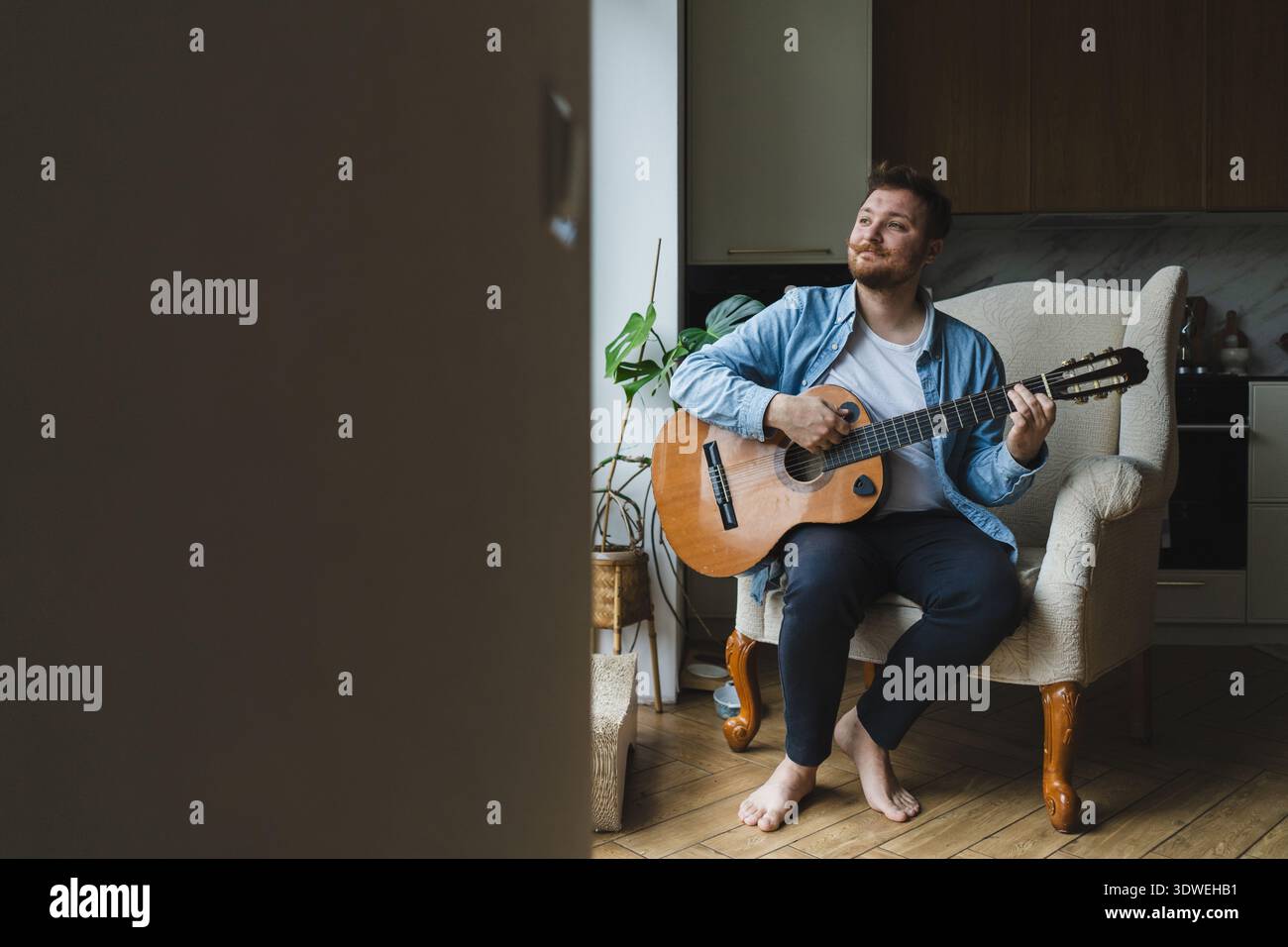 Un giovane uomo siede comodamente nel suo salotto, suonando una chitarra con concentrazione e passione. L'ambiente accogliente e la luce naturale creano un'atmosfera serena Foto Stock