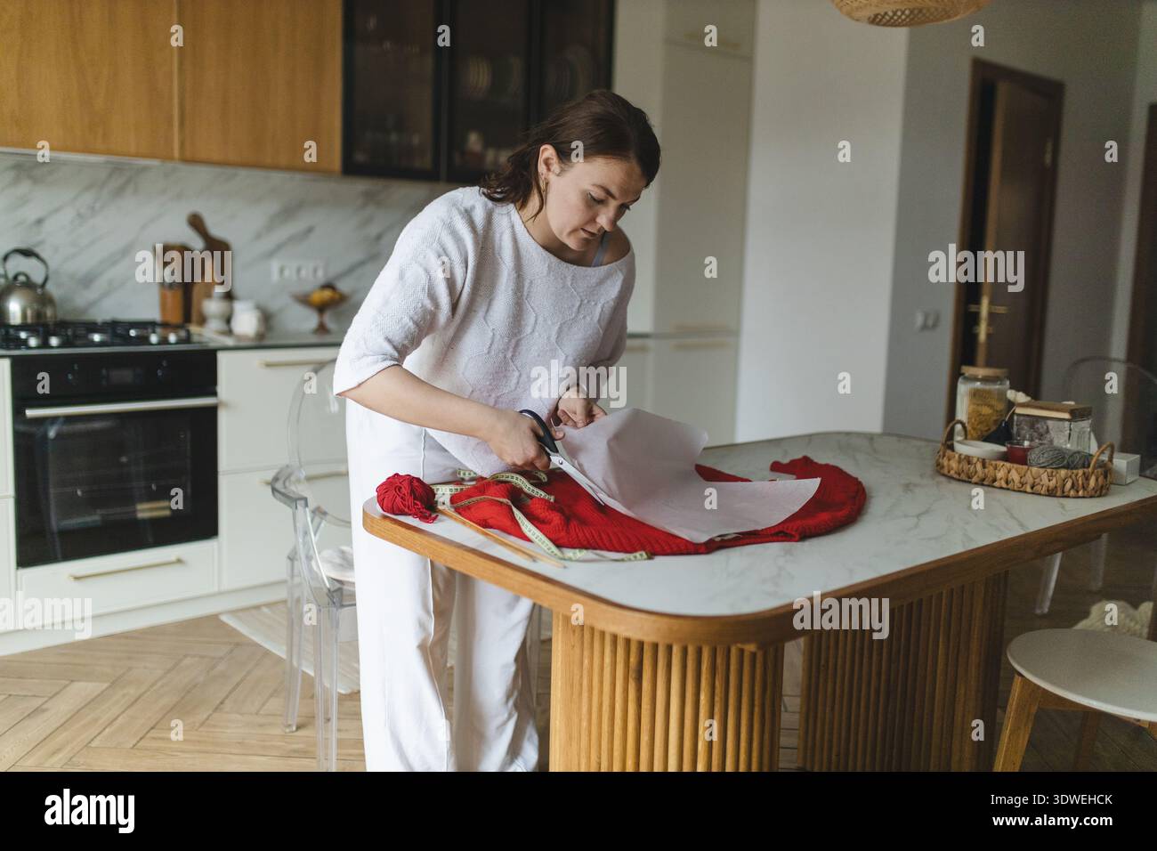 Una donna è vista in una cucina accogliente, impegnata a creare un maglione rosso. Taglia con cura la carta, circondata da filati e attrezzi, incarnando la passione per lui Foto Stock