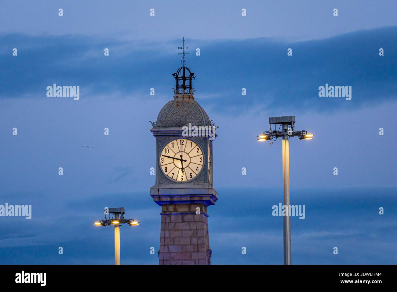 Una maestosa torre dell'orologio urbano si erge su di un cielo crepuscolo profondo, fiancheggiata da lampioni luminosi. L'architettura classica completa la struttura Foto Stock
