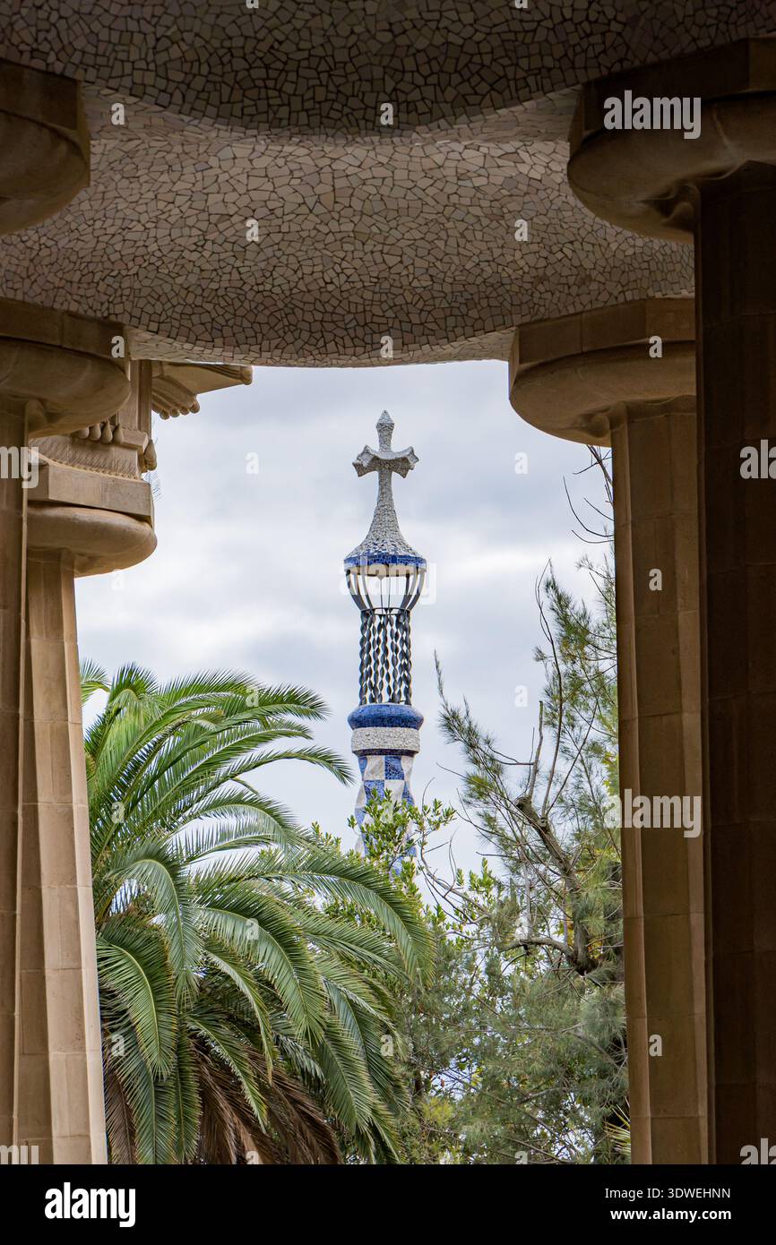Vista attraverso la terrazza colonnata di Park Guell, che mostra un pilastro coperto di mosaico con una croce in cima, circondato dalla vegetazione lussureggiante di Barcellona, Spagna Foto Stock