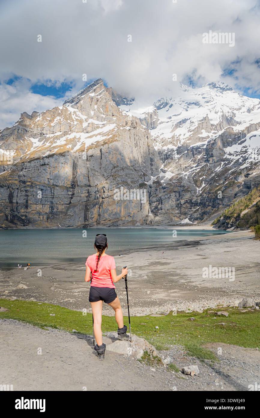 Un escursionista con un abbigliamento vivace si erge sullo splendido lago Oeschinen, annidato nelle Alpi svizzere. Le cime innevate e le foreste lussureggianti forniscono un pict Foto Stock