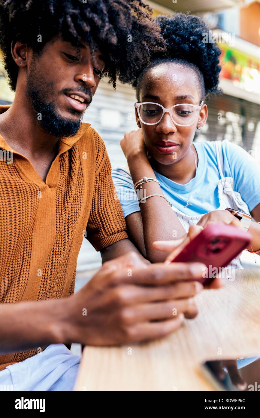 Una coppia si siede vicino a un tavolo, condividendo un momento di gioia guardando lo schermo di uno smartphone. Il loro comportamento rilassato e allegro indica un positi Foto Stock