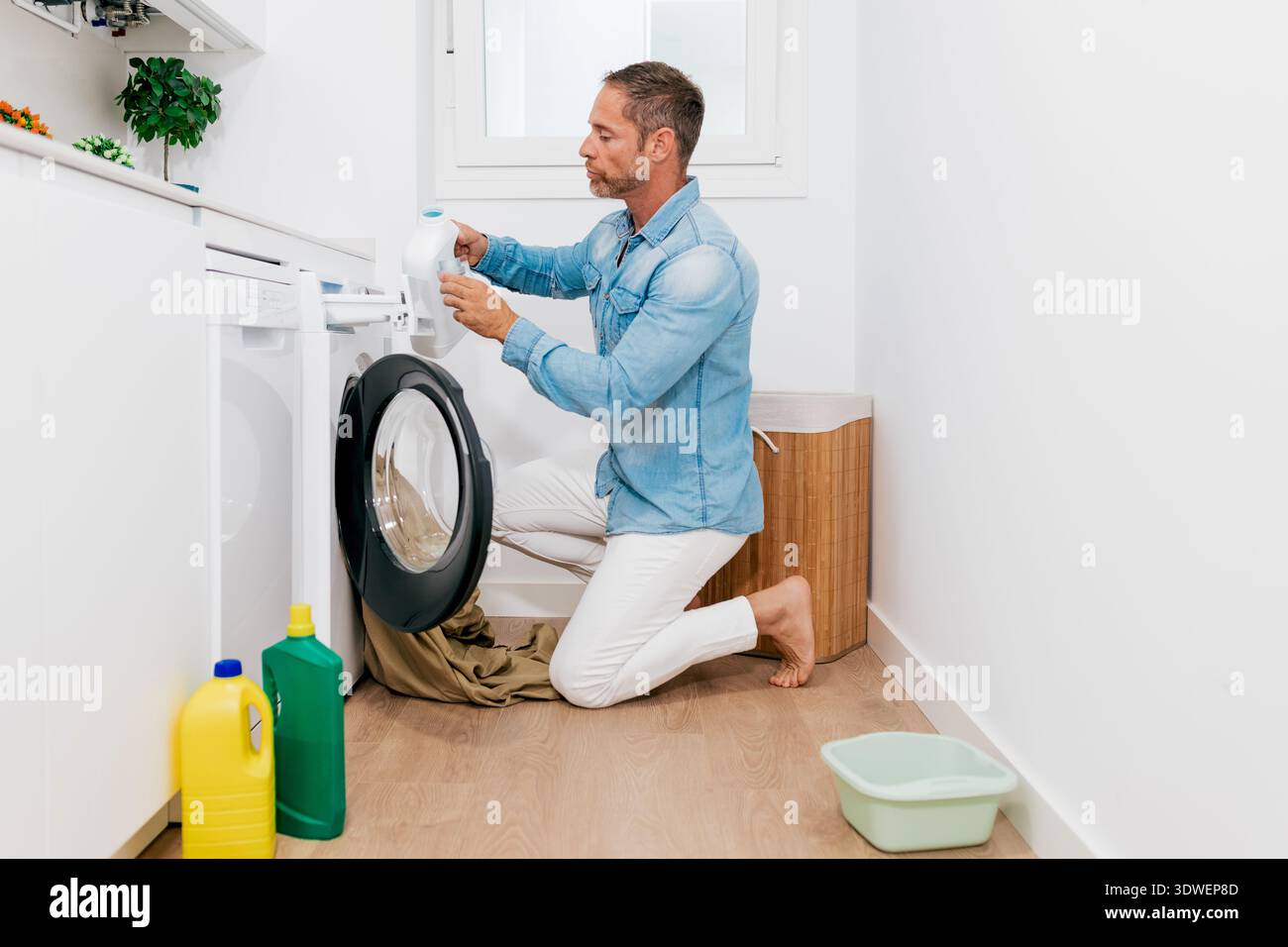 Un uomo si sta inginocchiando in una lavanderia luminosa e moderna, aggiungendo detersivo a una lavatrice a caricamento frontale. Si vedono vari materiali di pulizia, enfatizzini Foto Stock