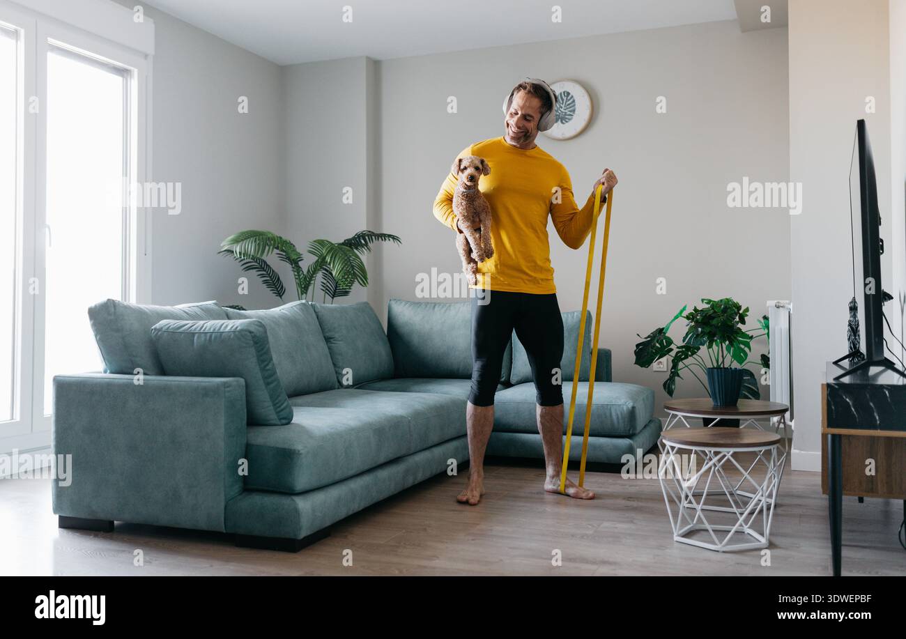 Un uomo con una camicia gialla si allena a casa usando una fascia di resistenza mentre tiene in mano un piccolo cane. Si trova a piedi nudi nel suo moderno soggiorno, circondato da Foto Stock