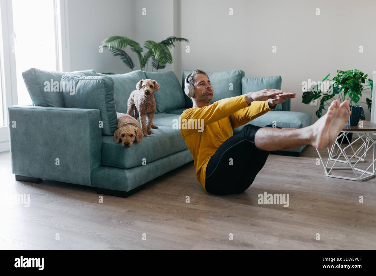 Un uomo si allena a casa, si siede sul pavimento indossando le cuffie. Due cani curiosamente lo guardano da un divano turchese, creando un c Foto Stock