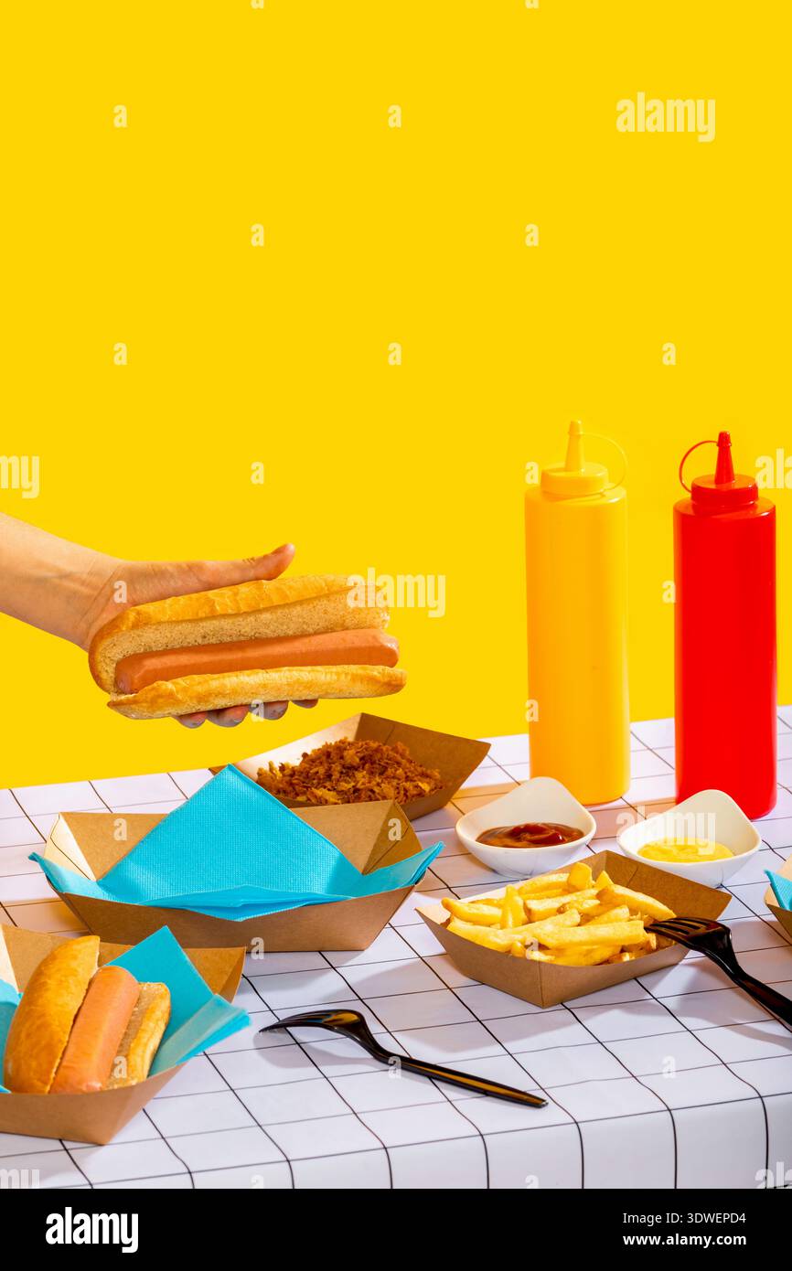 Un'immagine vibrante di un hot dog tenuto in mano su uno sfondo giallo brillante. Accompagnata da patatine fritte, salse e condimenti, questa scena cattura un clas Foto Stock
