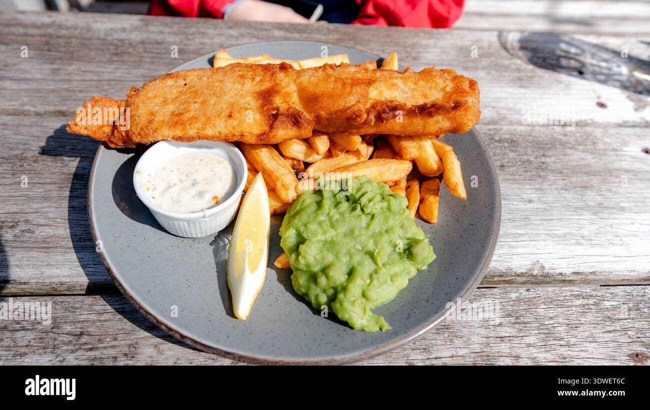Un piatto classico di fish and chips viene servito con piselli moschi, salsa di tartaro e un cuneo di limone su un rustico tavolo in legno, catturando l'essenza del Brit Foto Stock