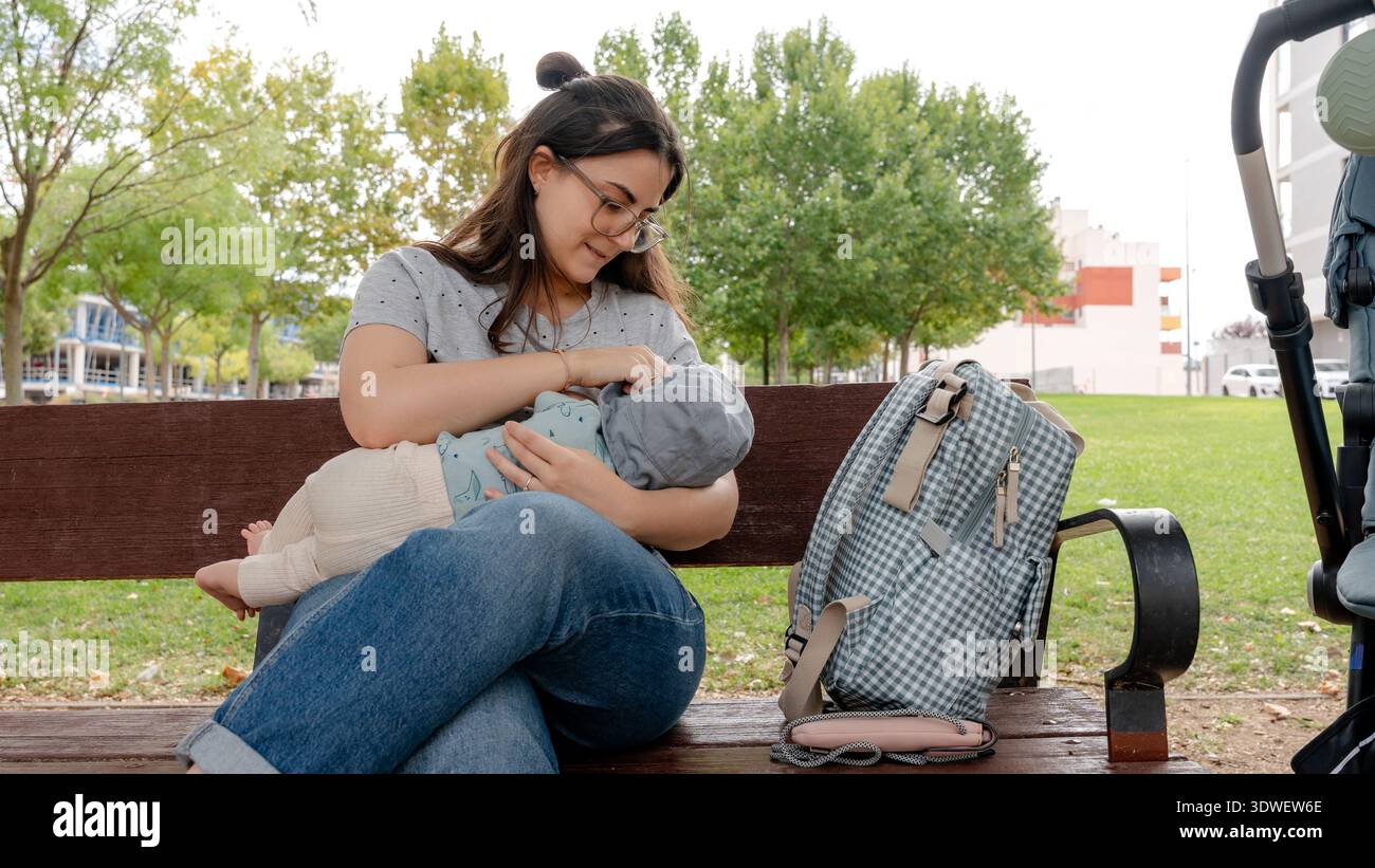 Una madre amorevole allatta il suo bambino su una panchina del parco, circondata dalla natura. La sua borsa e il passeggino nelle vicinanze completano questo sereno momento in famiglia in libertà Foto Stock
