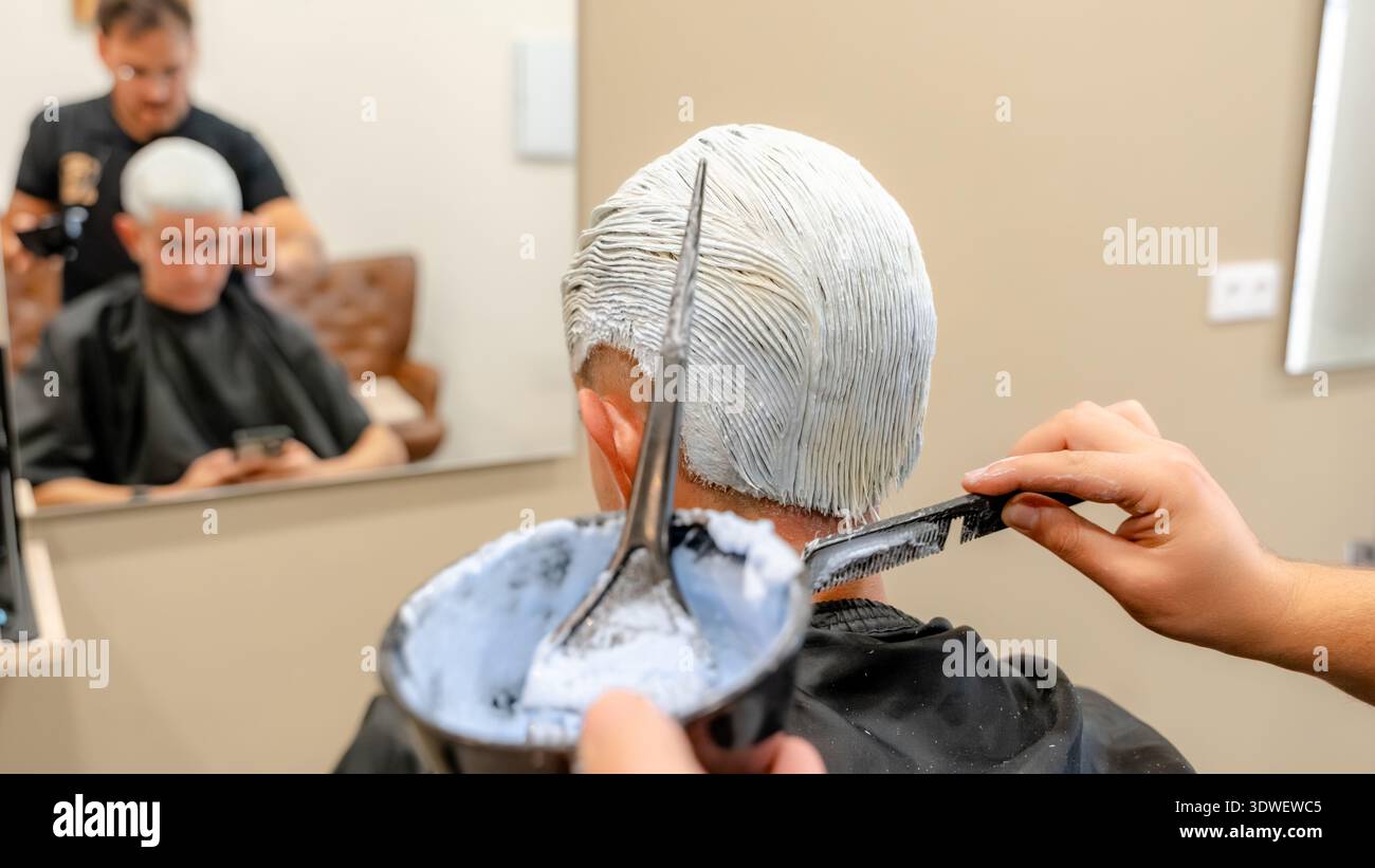 Un cliente in un barbiere sta facendo sbiancare i capelli, con uno stilista che applica meticolosamente il prodotto sbiancante. Il processo viene acquisito con gli strumenti Foto Stock