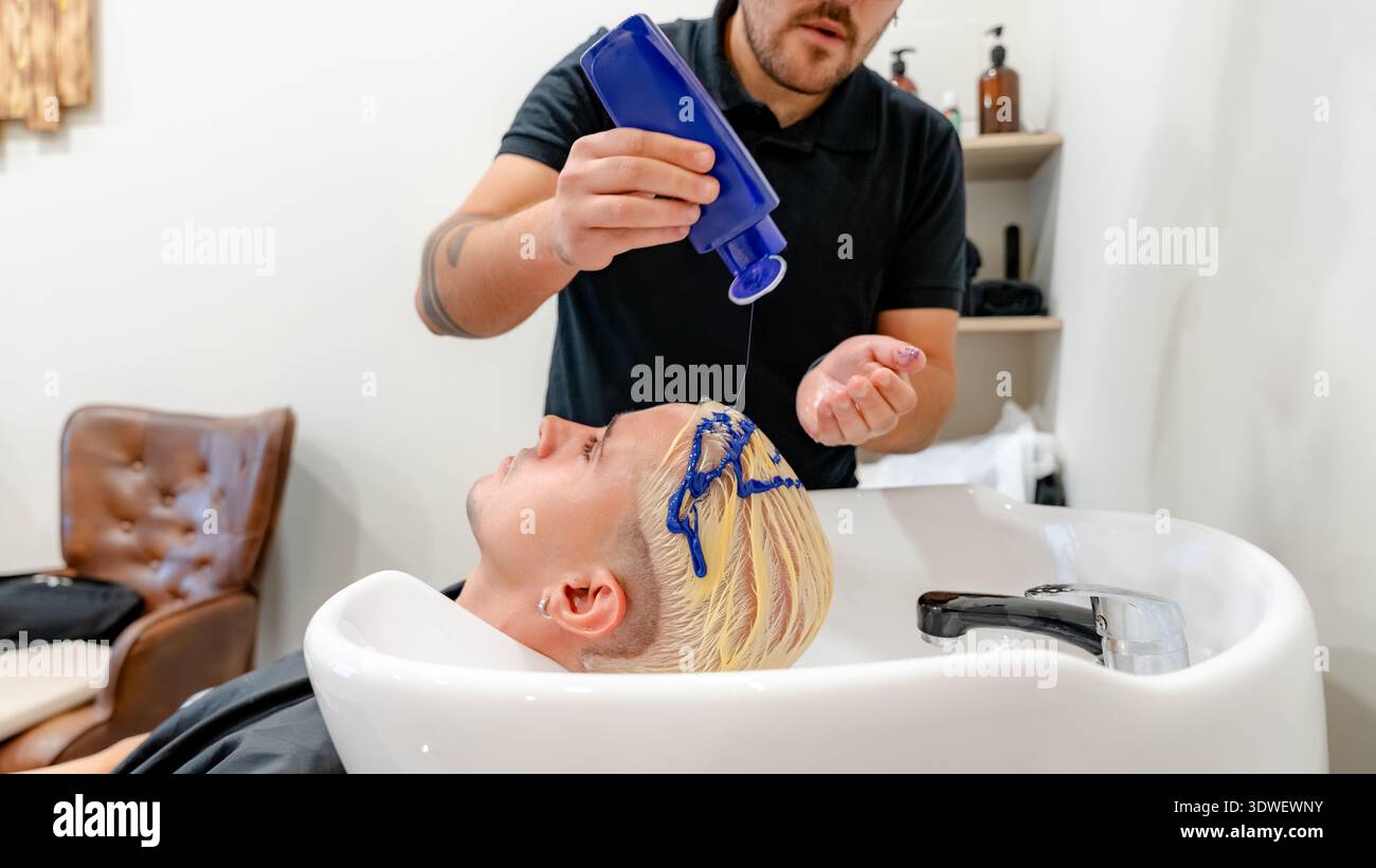 Un barbiere applica il toner blu ai capelli sbiancati di un cliente in un elegante barbiere. L'immagine acquisisce la tecnica precisa e gli strumenti moderni utilizzati nella Foto Stock