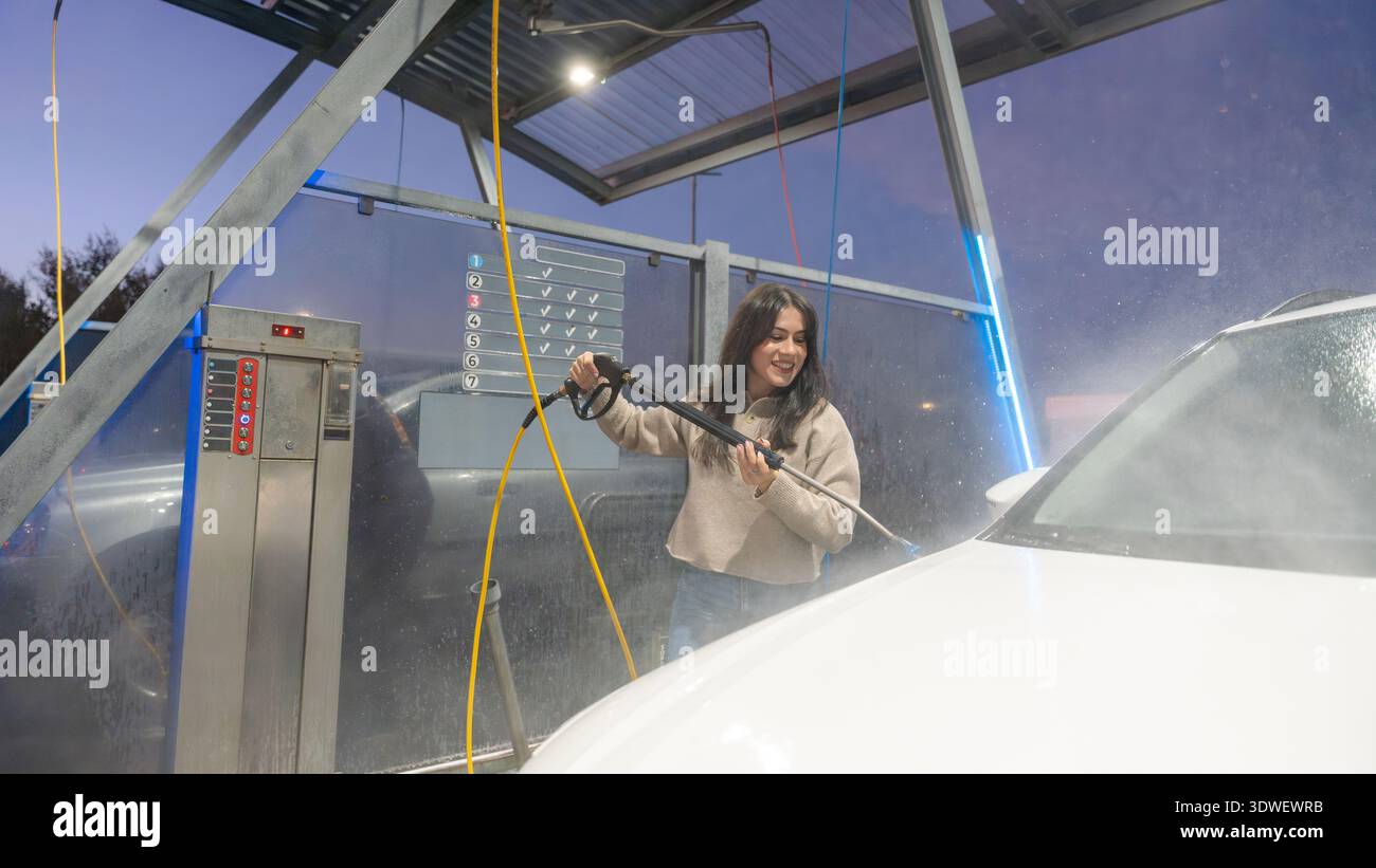Una donna sorride mentre usa un'idropulitrice ad alta pressione per pulire la sua auto in un autolavaggio self-service all'aperto. Il sole che tramonta proietta una serena luce soffusa sulla m Foto Stock