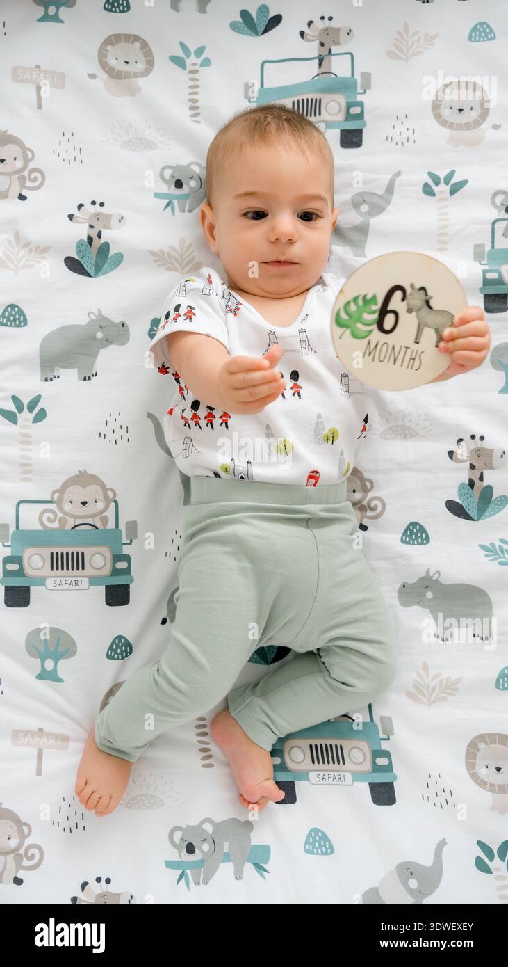 Un bambino di sei mesi detiene una carta miliare di sei mesi mentre sdraiato su una giocosa coperta a tema safari con illustrazioni di animali carini e un safari Foto Stock