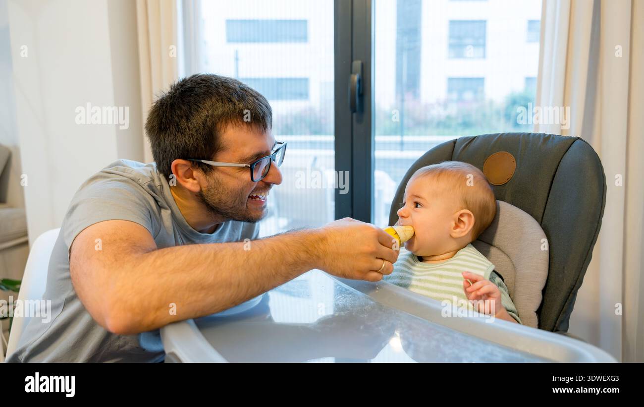 Un genitore affettuoso nutre al suo bambino una banana come parte dello svezzamento condotto dal bambino. Il bambino, seduto su un seggiolone, esplora il gusto e la consistenza del frutto, Foto Stock
