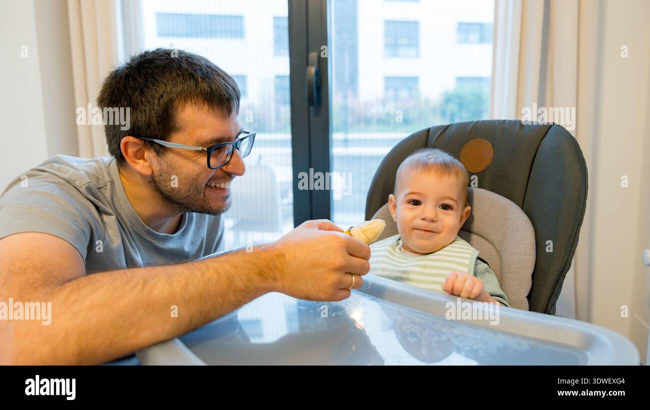 Un padre condivide con gioia una banana con il suo bambino durante un pasto in cucina. Il bambino, seduto su un seggiolone, sembra curioso ed entusiasta di tr Foto Stock