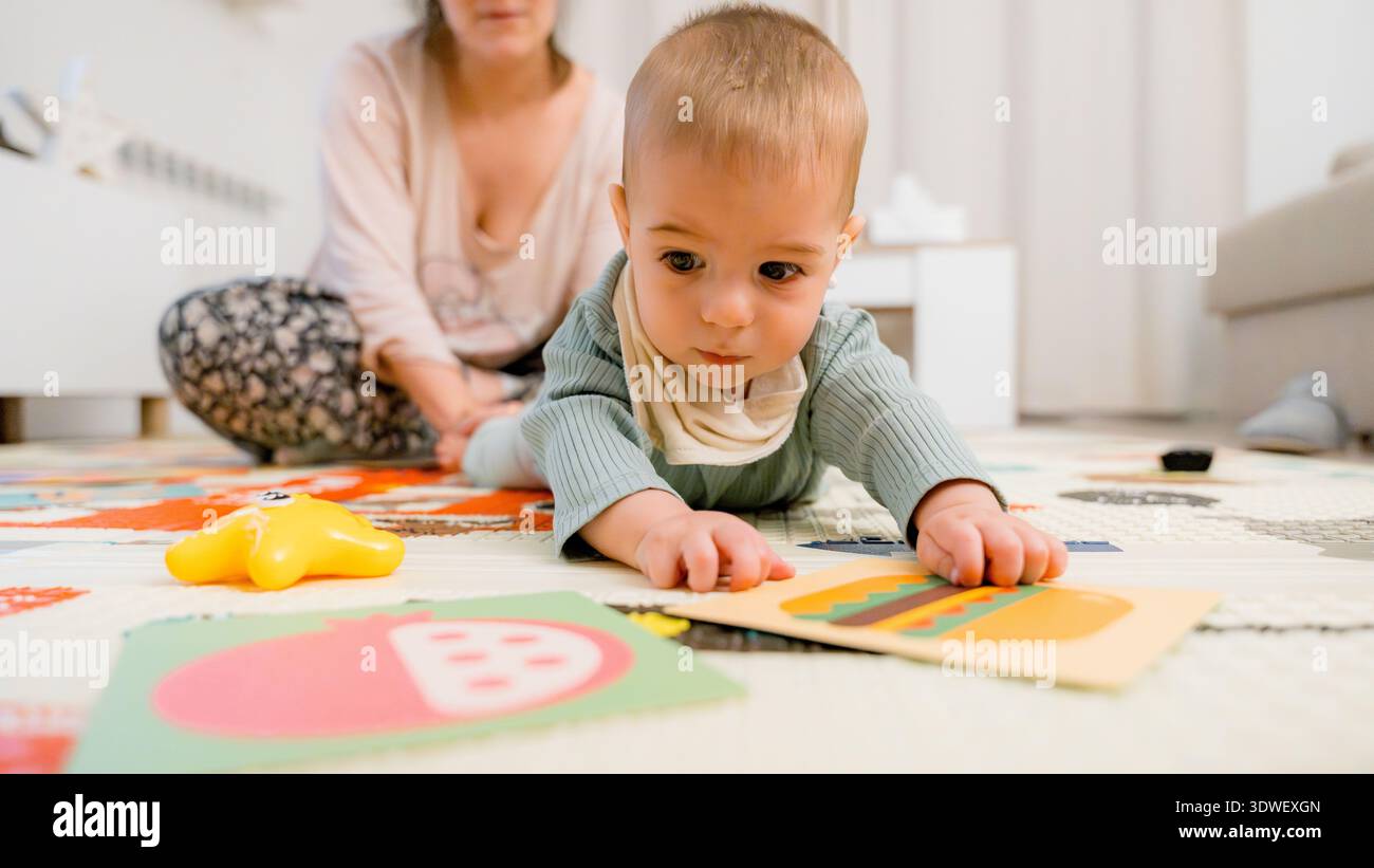 Un bambino curioso in un intimo vestito esplora le carte didattiche colorate su un morbido tappetino, promuovendo l'apprendimento precoce, con un attento attento operatore sanitario che guarda Foto Stock