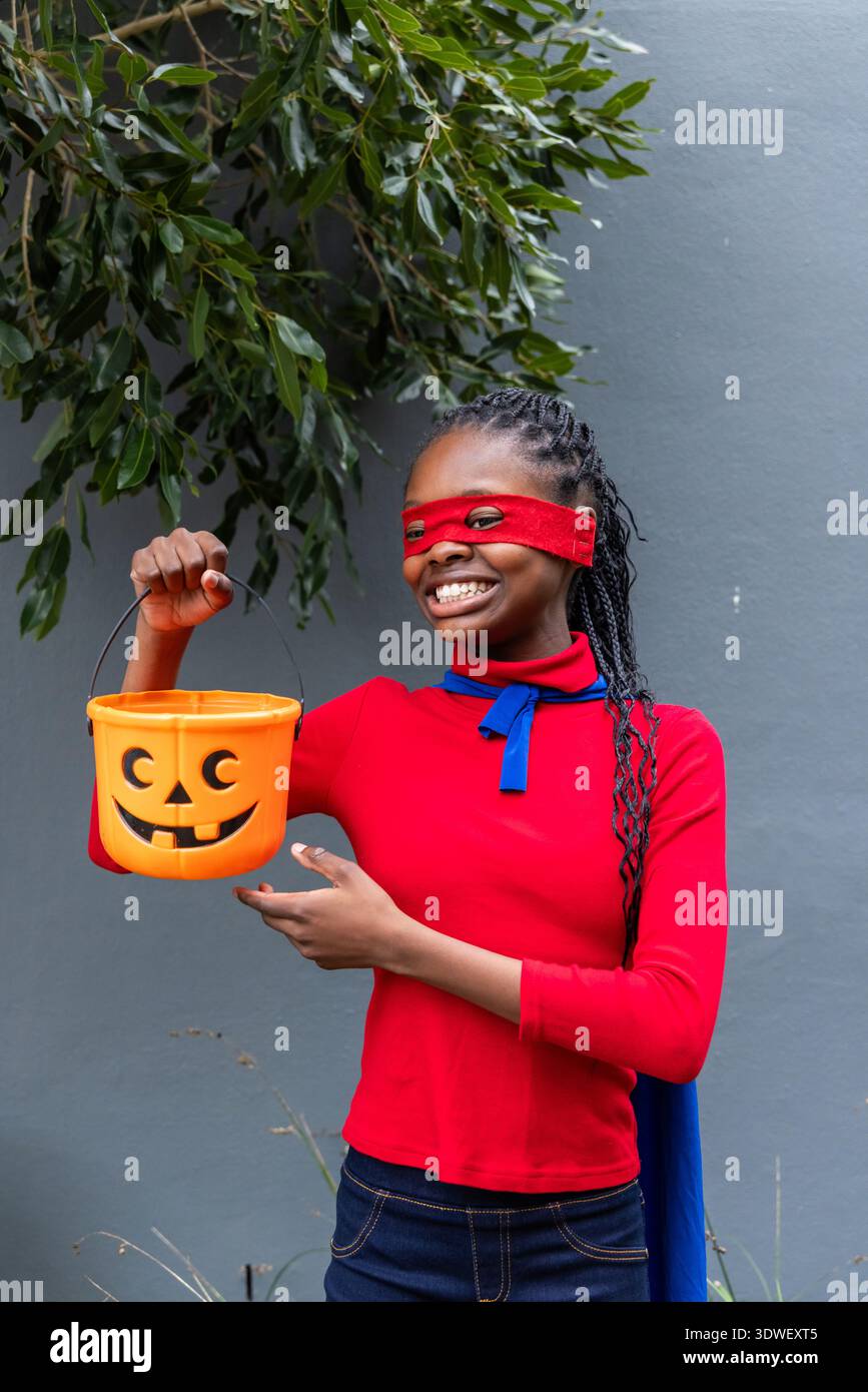 Ragazza afro-americana che posa sul muro grigio, indossa una maschera rossa, mantello blu, tenendo in mano il secchio arancione Foto Stock