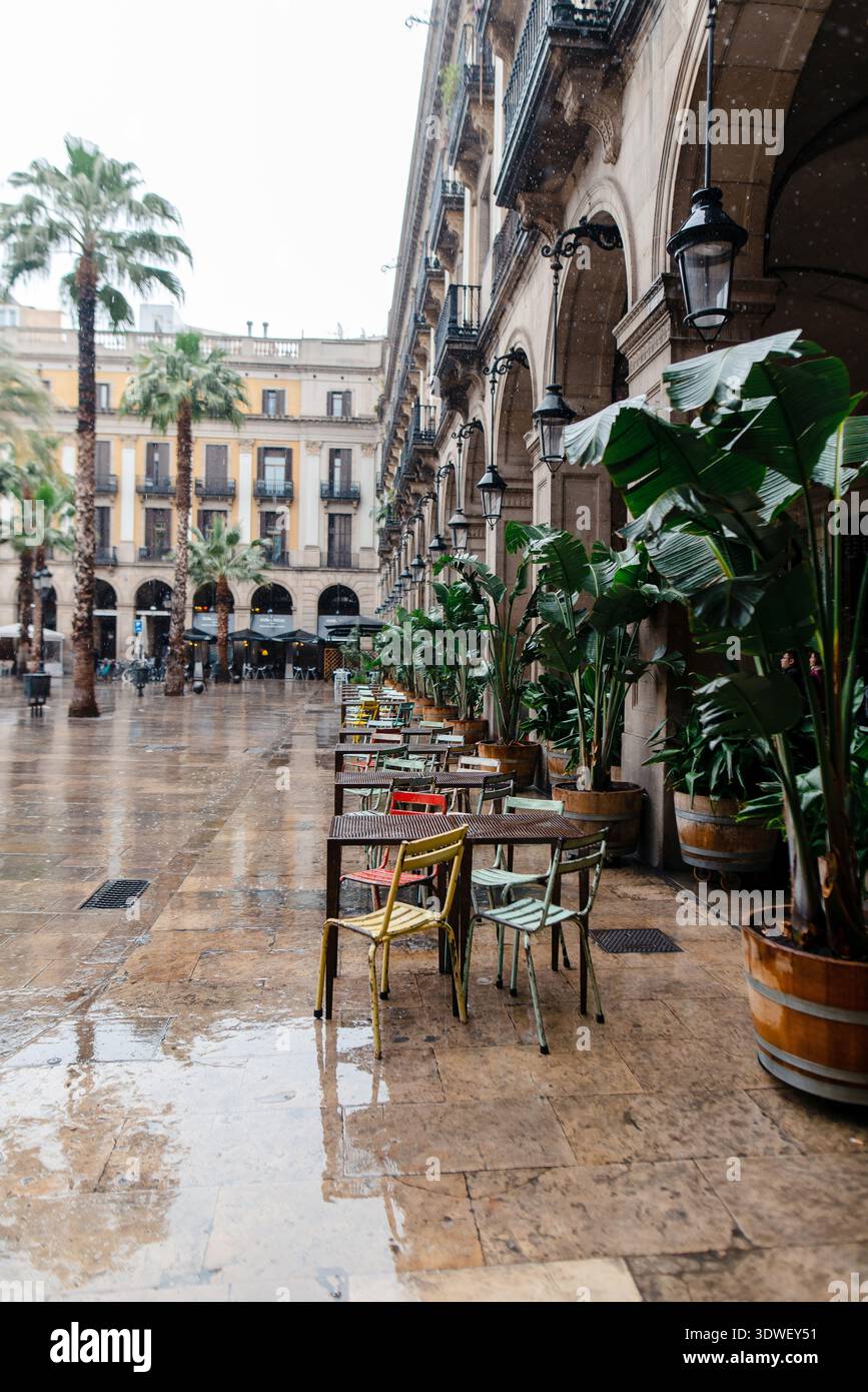 Area vuota con posti a sedere all'aperto di un caffè a Barcellona, Spagna, con pavimentazione bagnata che riflette il cielo coperto e l'architettura circostante Foto Stock