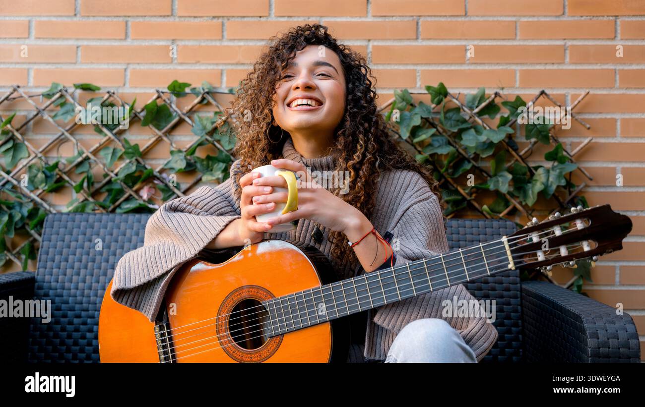 Una giovane donna con i capelli ricci si gode un momento di relax, tenendo una tazza mentre si siede con una chitarra. Trasuda calma e soddisfazione in un ambiente accogliente Foto Stock