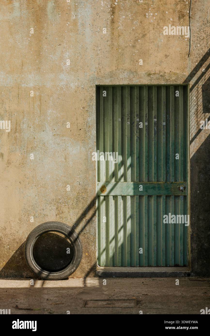 Scena illuminata dal sole con una porta in metallo arrugginito e una vecchia ruota. Le pareti testurizzate e le ombre aggiungono elementi di contrasto e profondità a questa architettura urbana Foto Stock