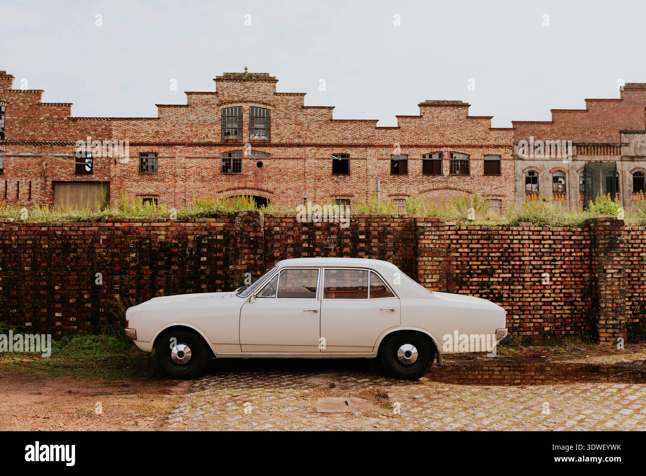Una classica auto bianca è parcheggiata di fronte a un edificio industriale in mattoni abbandonato, creando una scena nostalgica. La strada acciottolata e i mattoni rossi Foto Stock