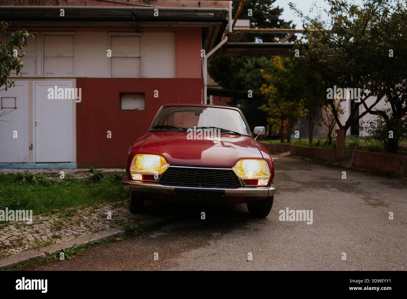 Una classica auto rossa d'epoca è parcheggiata in una strada tranquilla e stretta al crepuscolo. I fari dell'auto sono accesi, illuminando la pavimentazione in ciottoli e il suo carattere Foto Stock