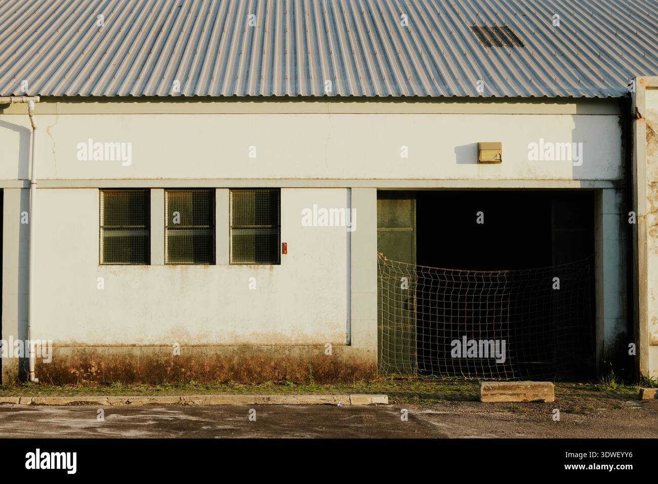 Un vecchio magazzino con una facciata resistente agli agenti atmosferici e tetto in metallo ondulato sotto la calda luce del sole. L'ingresso, parzialmente coperto da una rete, aggiunge un ingresso industriale Foto Stock