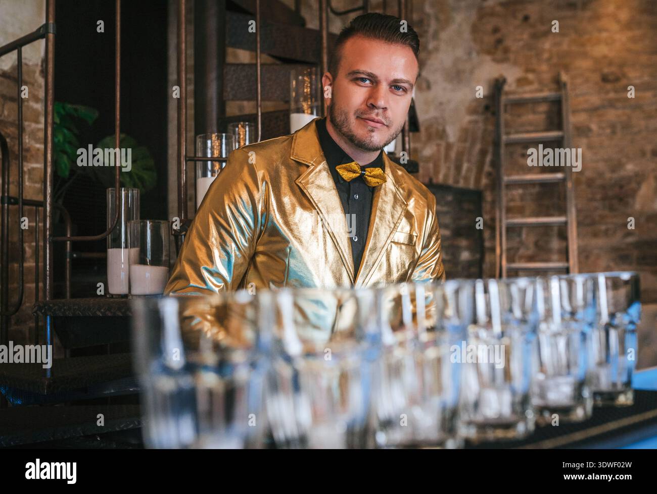 Un barista in una vivace giacca dorata inizia a preparare cocktail, concentrandosi su una fila di bicchieri vuoti pronti per l'inizio di una serata vivace, ambientando un Foto Stock