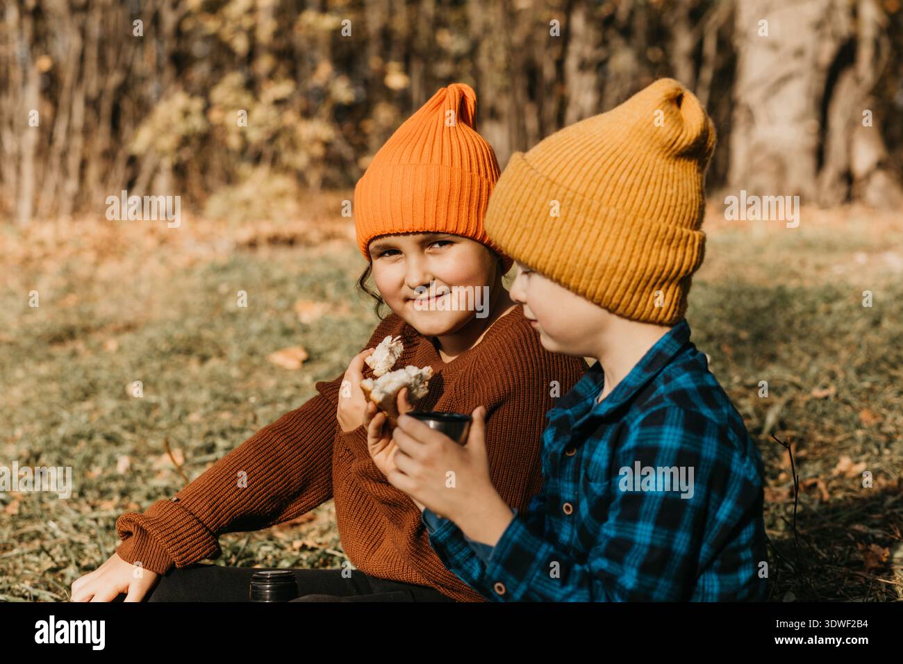 Due bambini che indossano colorati cappelli a maglia sono seduti in una foresta durante l'autunno. Appaiono contenti e gioiosi, gustando spuntini mentre sono circondati da Foto Stock