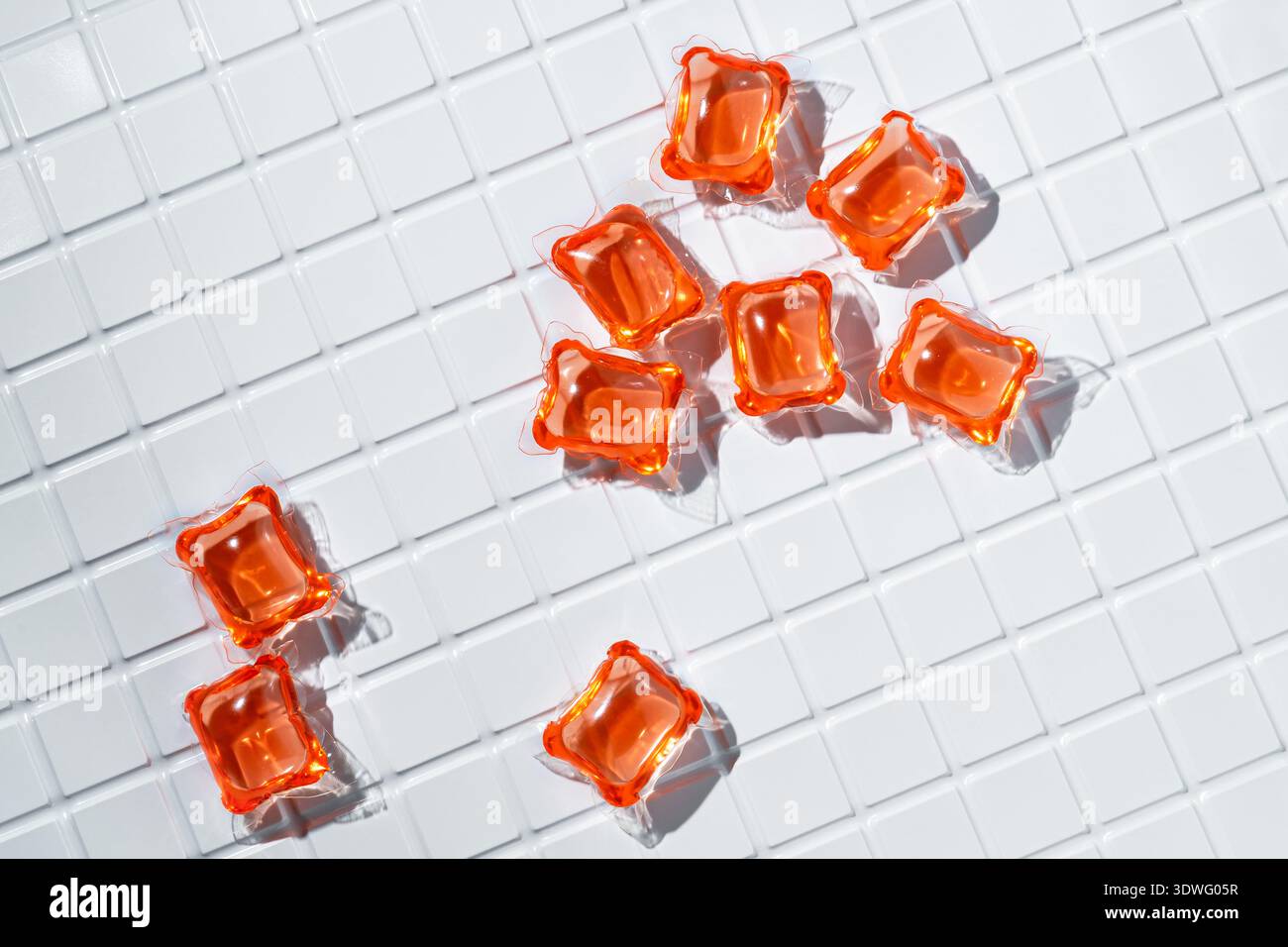 Capsule per bucato di colore arancione brillante disposte su una superficie piastrellata bianca. L'esterno lucido delle cialde riflette la luce, sottolineandone i colori vivaci e. Foto Stock