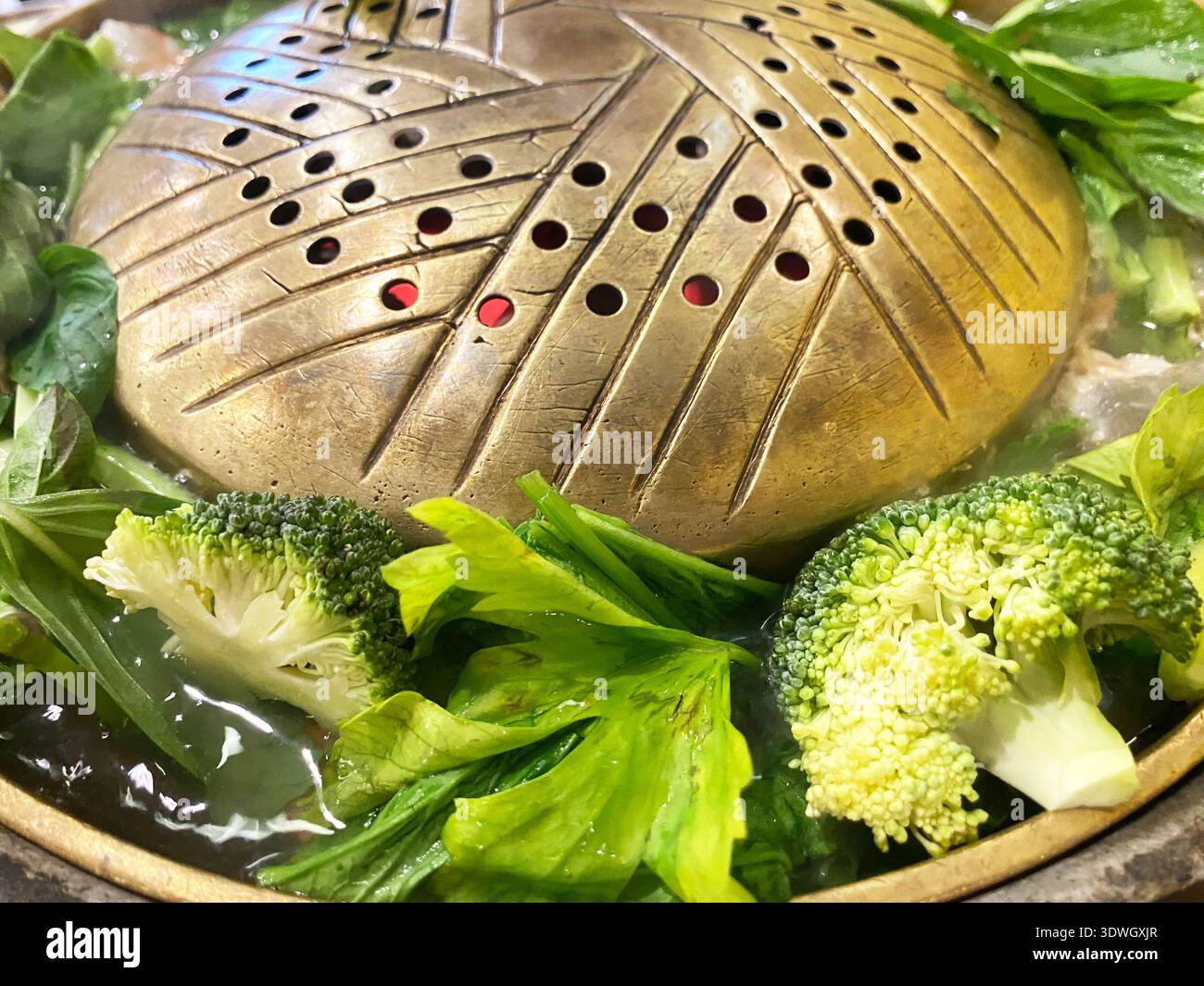 Pentola calda metallizzata ripiena di brodo fumante e verdure fresche come broccoli e verdure a foglia verde, in un vivace ambiente ristorante Foto Stock