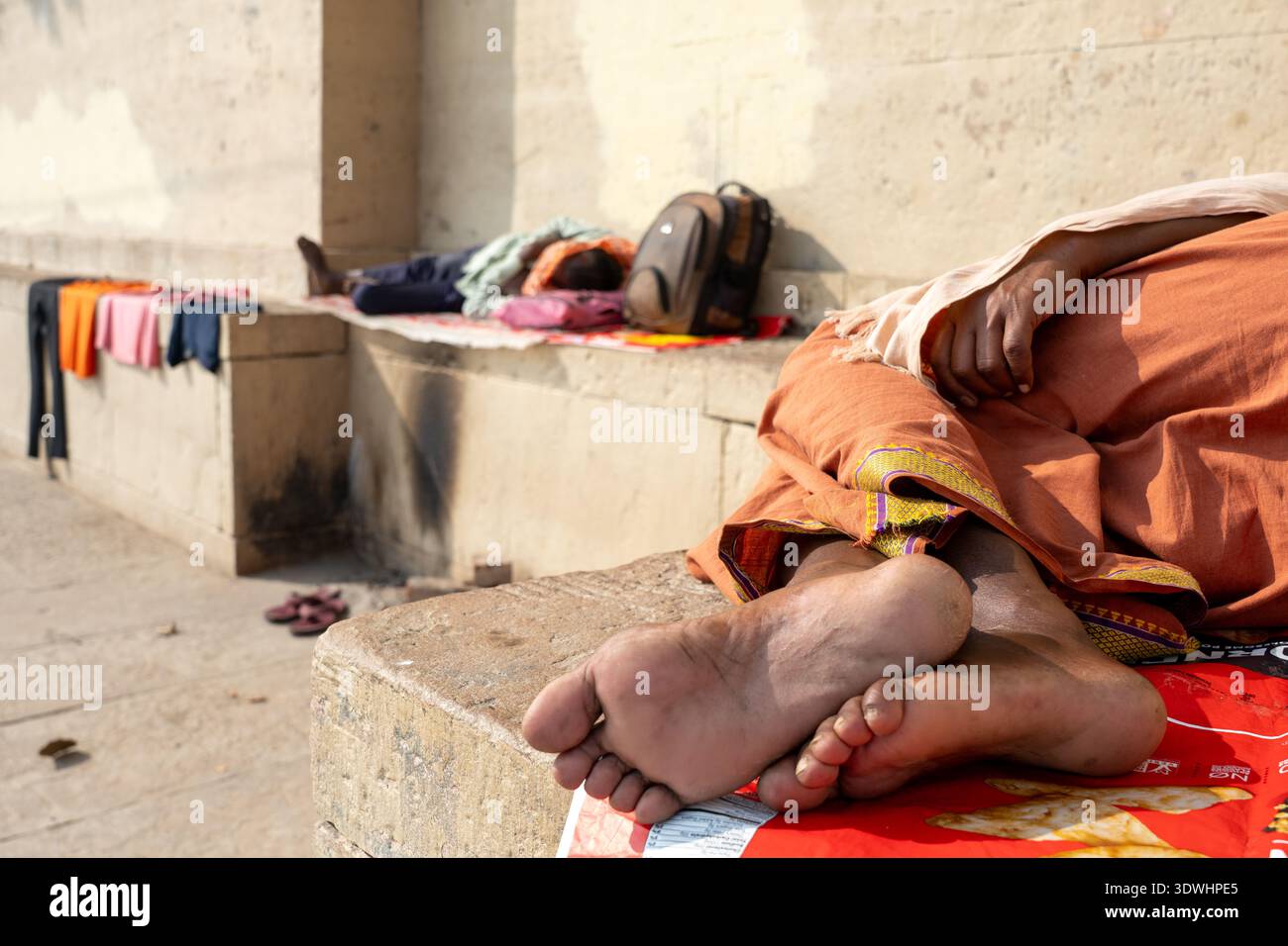 Due persone riposano su panchine di pietra a Varanasi, in India, con vestiti e borse accanto alla luce del sole. Foto Stock