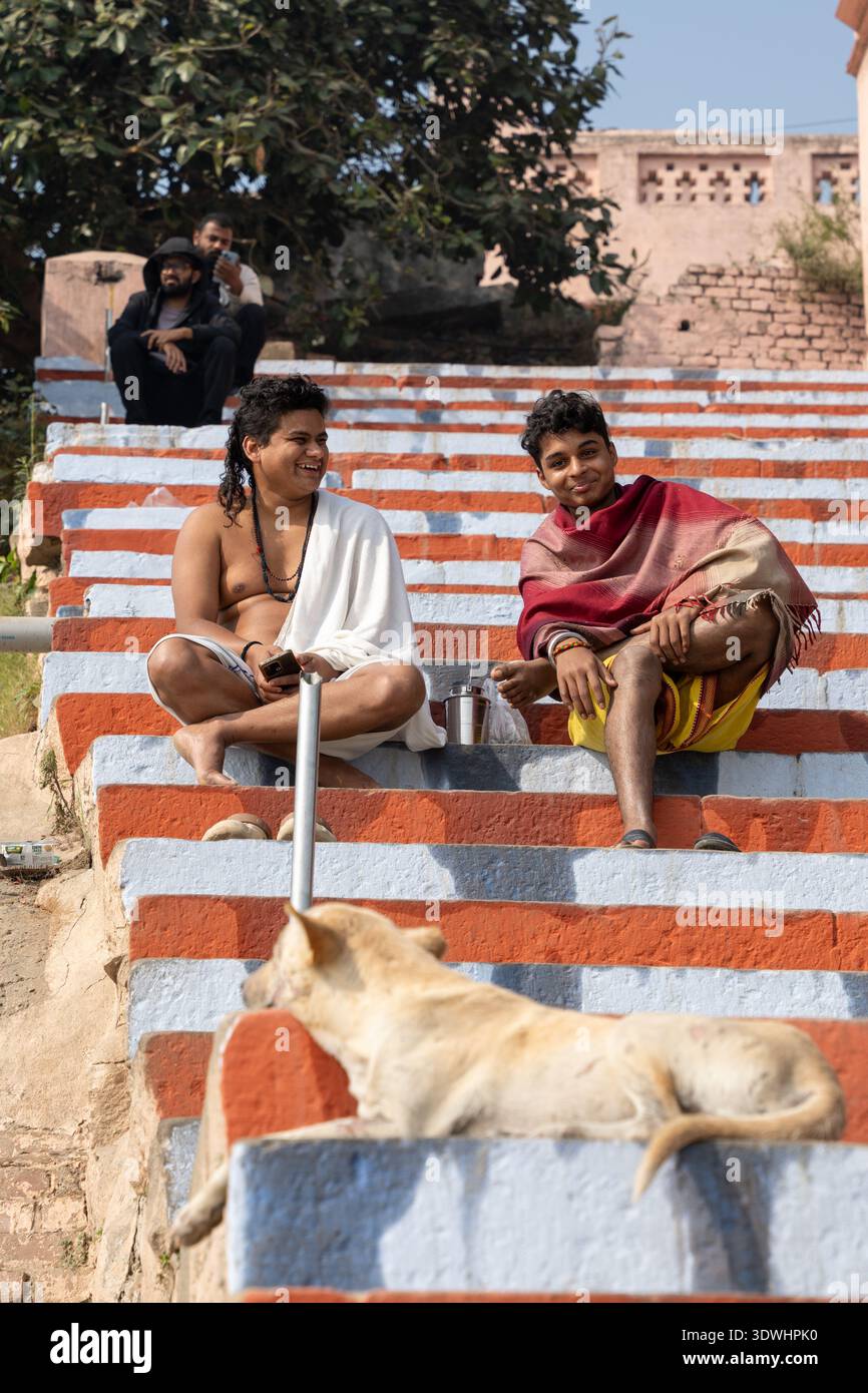 Due giovani uomini si rilassano su gradini dipinti con un cane a Varanasi, India, godendosi una giornata di sole all'aperto. Foto Stock
