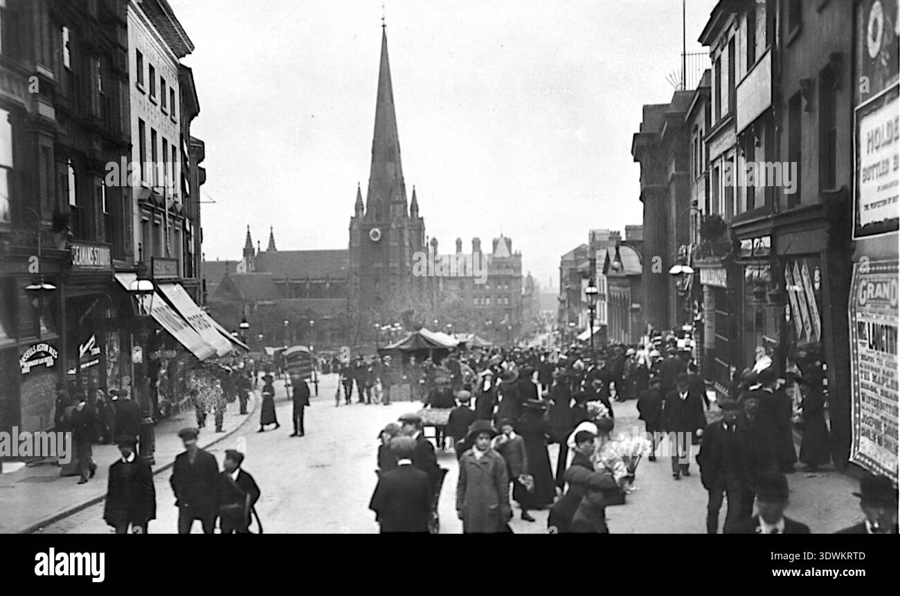 Il Bull Ring è pieno di energia all'inizio del XX secolo: Bancarelle del mercato, commercianti e dense folle riempiono la strada, incorniciata da alti edifici commerciali. Una guglia della chiesa sorge al centro della vista, ancorando la scena urbana. Uomini in cappello, donne e bambini si intrecciano nel vivace mercato che ha definito il cuore storico di Birmingham. Da una cartolina originale pubblicata dalla Rotary Photographic Company, intorno al 1906, Foto Stock