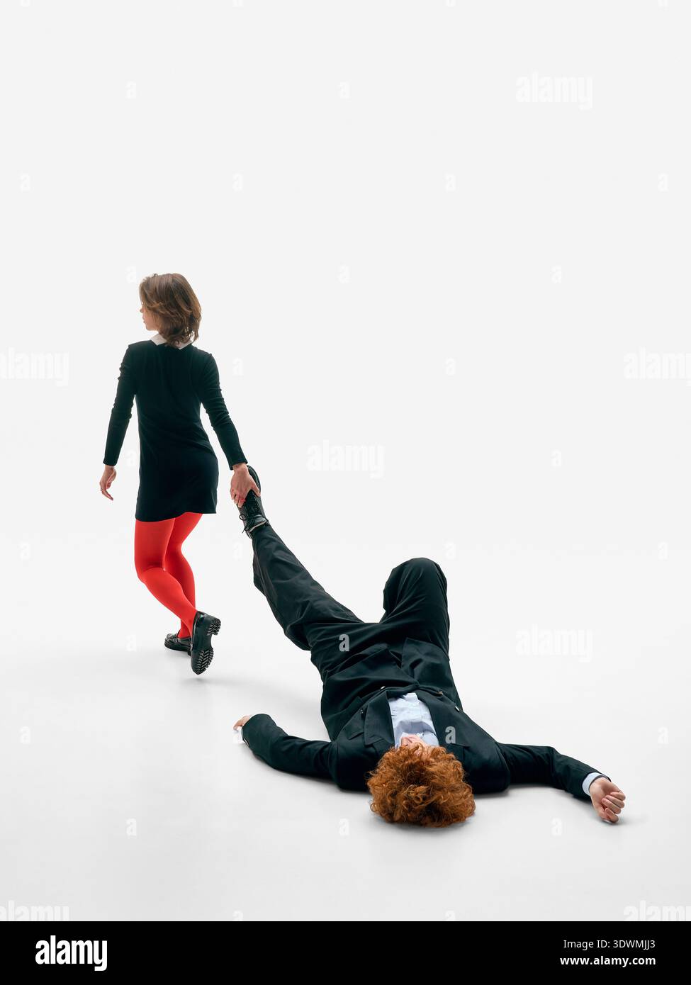 Donna che trascina uomo per gamba sul pavimento dello studio su sfondo bianco in abiti formali neri. Foto Stock
