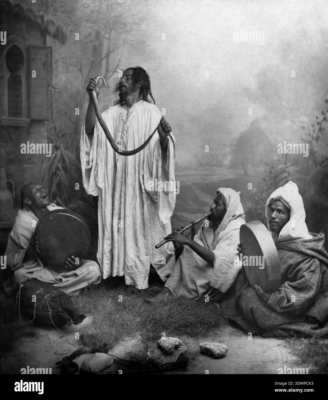 Questa fotografia antica della fine degli anni '1800 cattura un gruppo di incantatori di serpenti in Marocco, probabilmente Tangeri. Un uomo sta in piedi in modo prominente, tenendo un serpente in alto, mentre altri tre sono seduti, suonando un flauto e due tamburi a mano, creando una performance ritmica e affascinante. Foto Stock