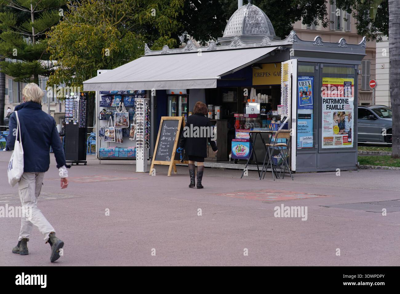 Classico chiosco di notizie grigio con riviste, cartoline e poster a Nizza, in Francia, una donna al banco e pedoni che passano sotto le palme. Foto Stock