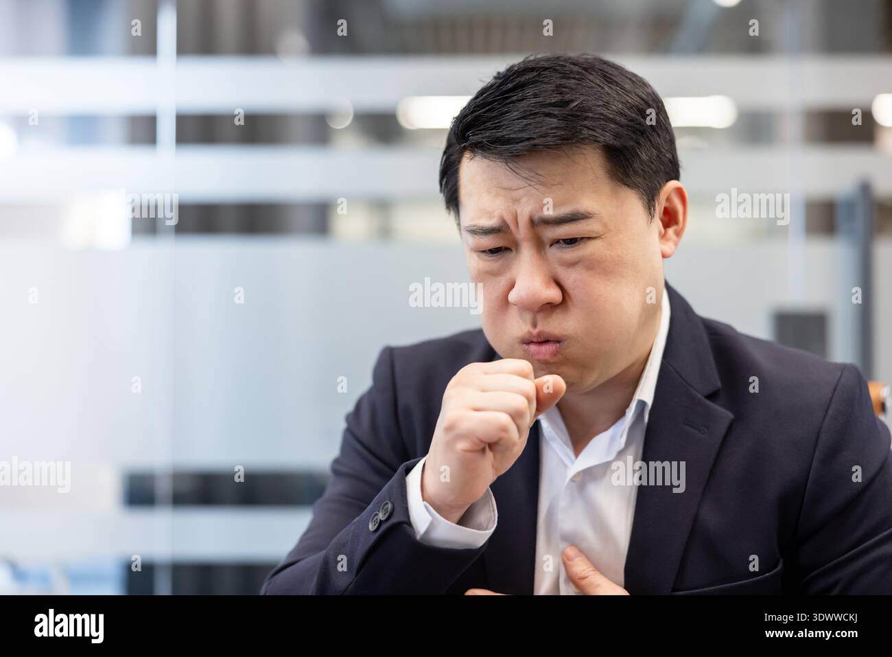 Uomo d'affari asiatico malato che soffre di una tosse dolorosa con un pugno vicino alla bocca e la mano sul petto, indicando disagio e malattia mentre lavora in un ufficio moderno Foto Stock