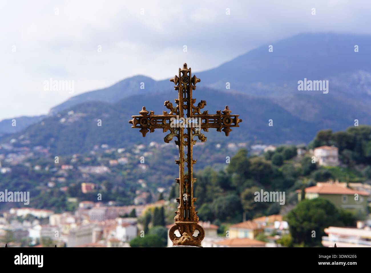 Croce ornata d'oro in cima al tetto contro un villaggio montagnoso e lo sfondo del cielo nuvoloso.Mentone, Francia Foto Stock