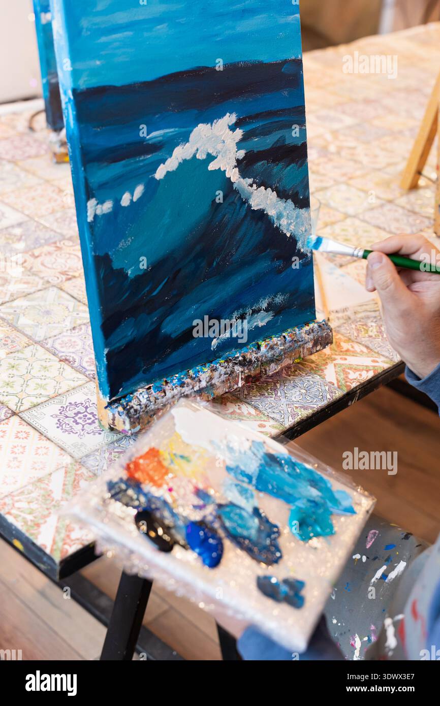 Un artista si concentra sulla pittura delle onde in sfumature di blu su tela. Tengono un pennello in una mano e una tavolozza piena di vari colori nell'altra, Foto Stock