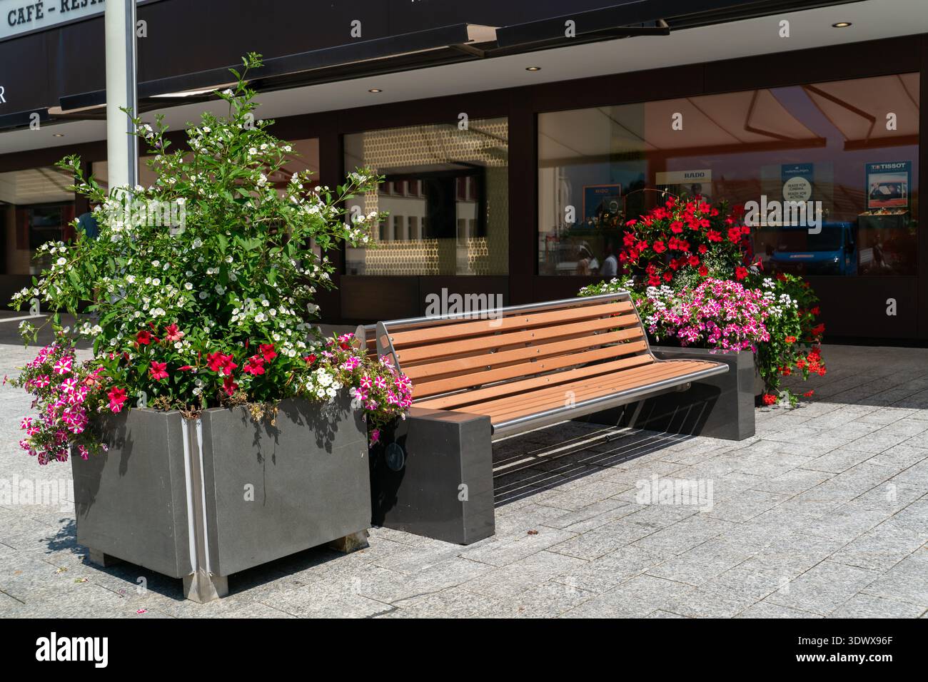 Panchina pubblica con spranghe in legno e struttura metallica fiancheggiata da fioriere decorative piene di fiori, esposta a Vaduz, Liechtenstein, che simboleggia l'urba Foto Stock