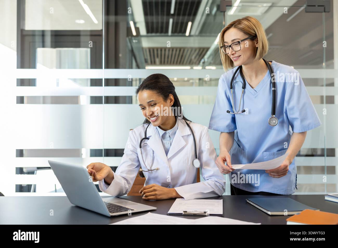 Due professionisti del settore medico multietnico sorridenti, un medico e un'infermiera, discutono dei dati dei pazienti e collaborano mentre guardano un portatile in uno studio ospedaliero Foto Stock